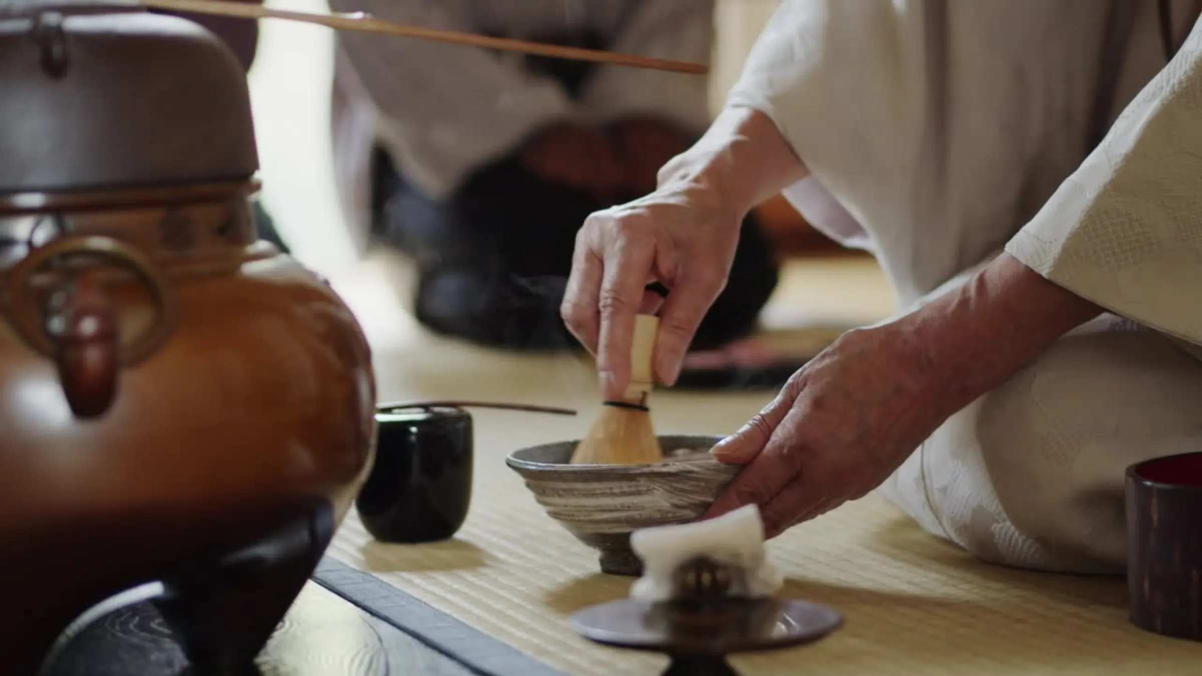 Whisking matcha in a chawan — steam rising beside the kettle on tatami